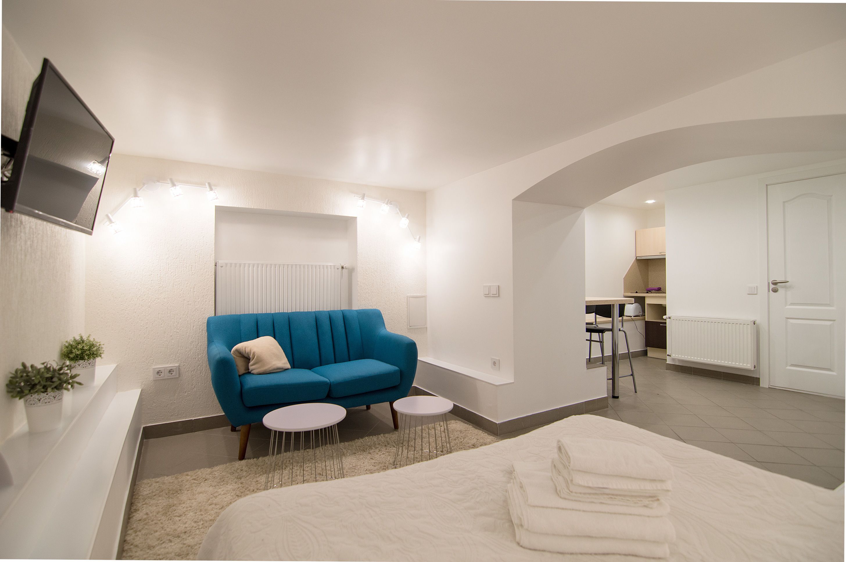 Moderni hotellihuone, jossa on sininen sohva, sänky, kaksi pientä pyöreää pöytää ja seinälle asennettu televisio.