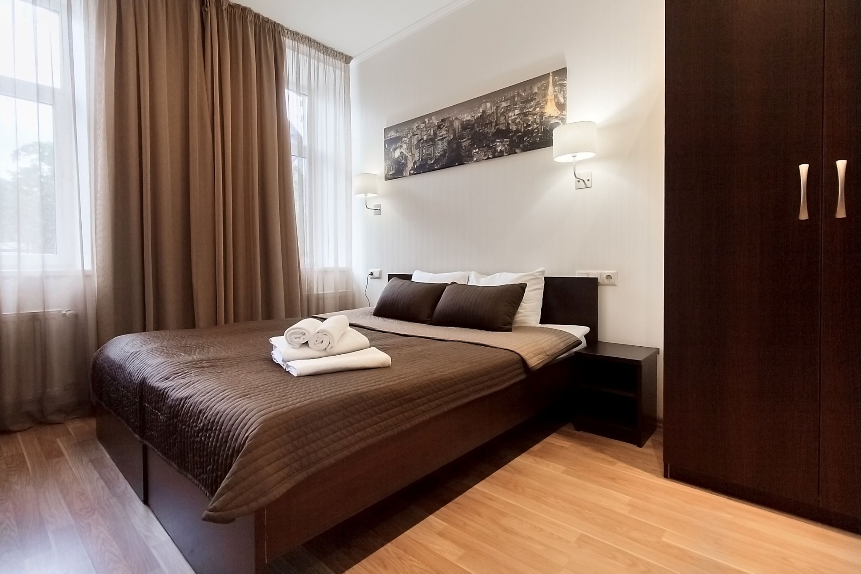 Quarto de hotel com uma cama grande, uma mesa de cabeceira, um guarda-roupa e uma pintura com vista para a cidade.