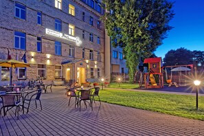 An outdoor patio area with tables and chairs, a children's play structure, and a building with a sign reading "Aparthotel Viesnica."
