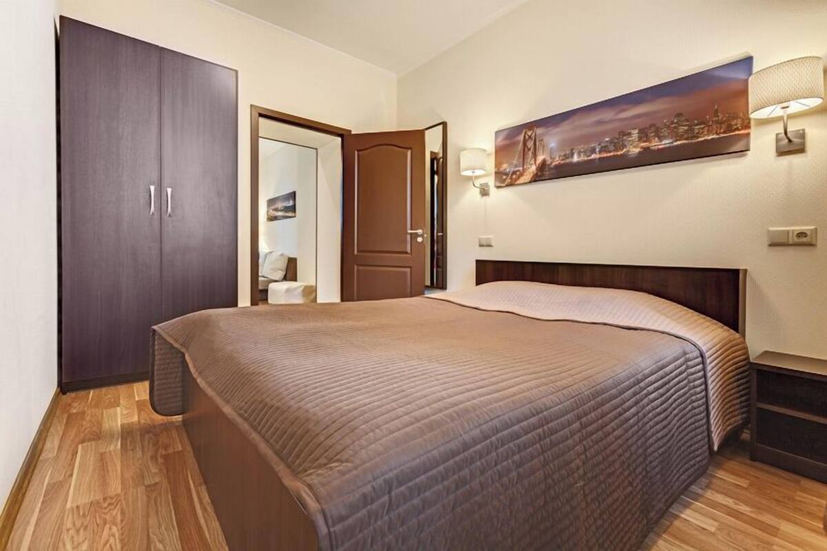 A bedroom with a bed, a wardrobe, a door, a nightstand, a lamp, and a wall-mounted picture.