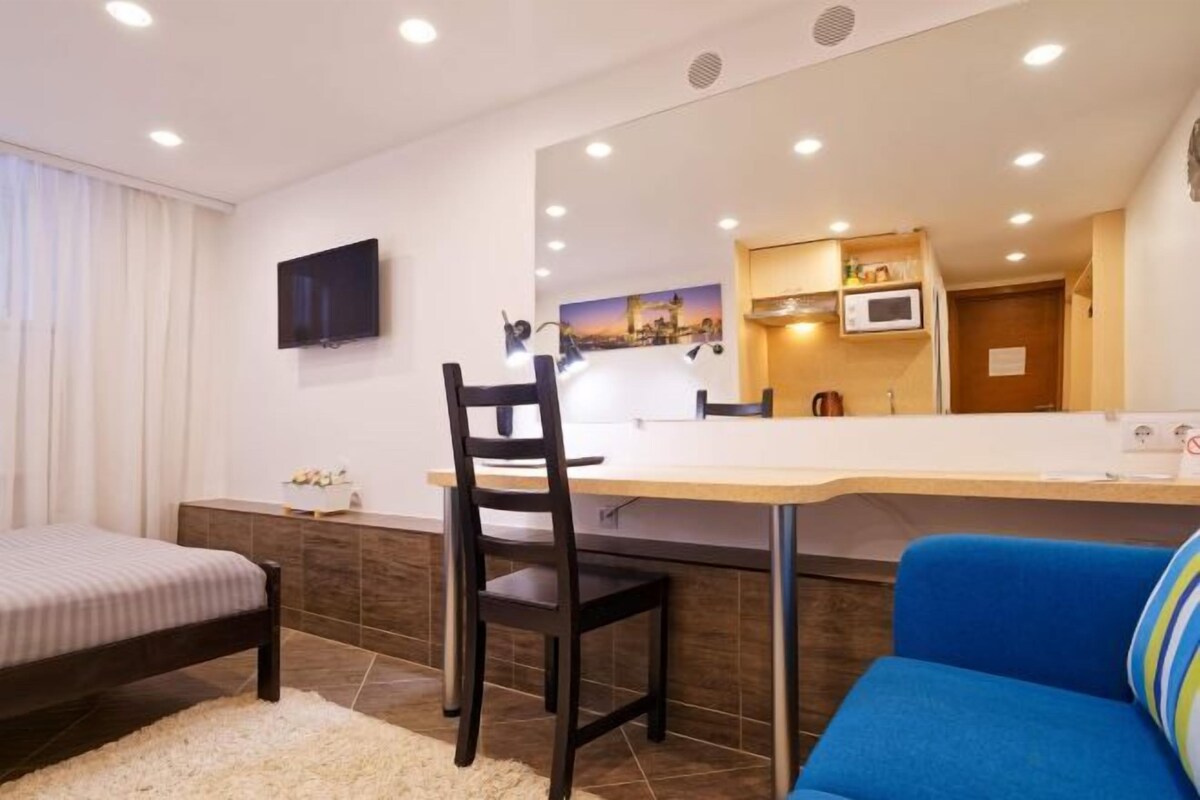 Een slaapkamer met een bed, een bureau, een stoel, een blauwe bank en een spiegel die een kitchenette weerkaatst.