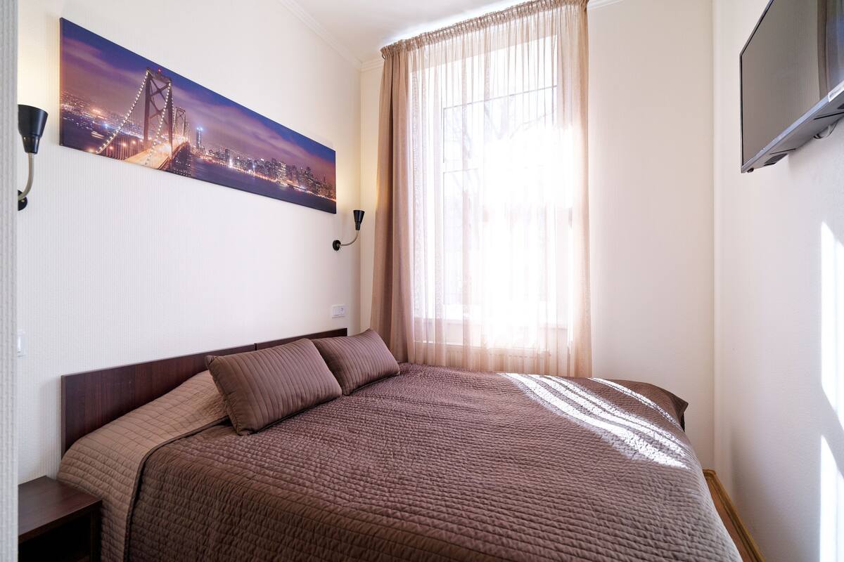 A neatly made bed with a brown quilt, a cityscape painting on the wall, a window with sheer curtains, and a mounted TV.