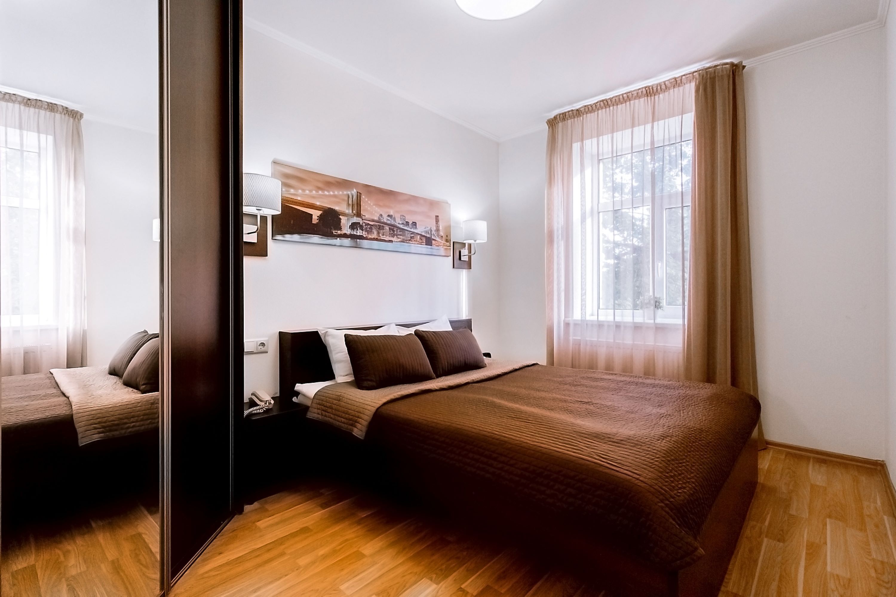 A hotel room with a large bed, a nightstand, a lamp, a painting on the wall, and a window with curtains.