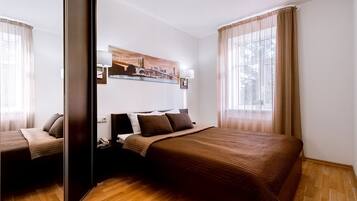 A hotel room with a large bed, a nightstand, a lamp, a painting on the wall, and a window with curtains.