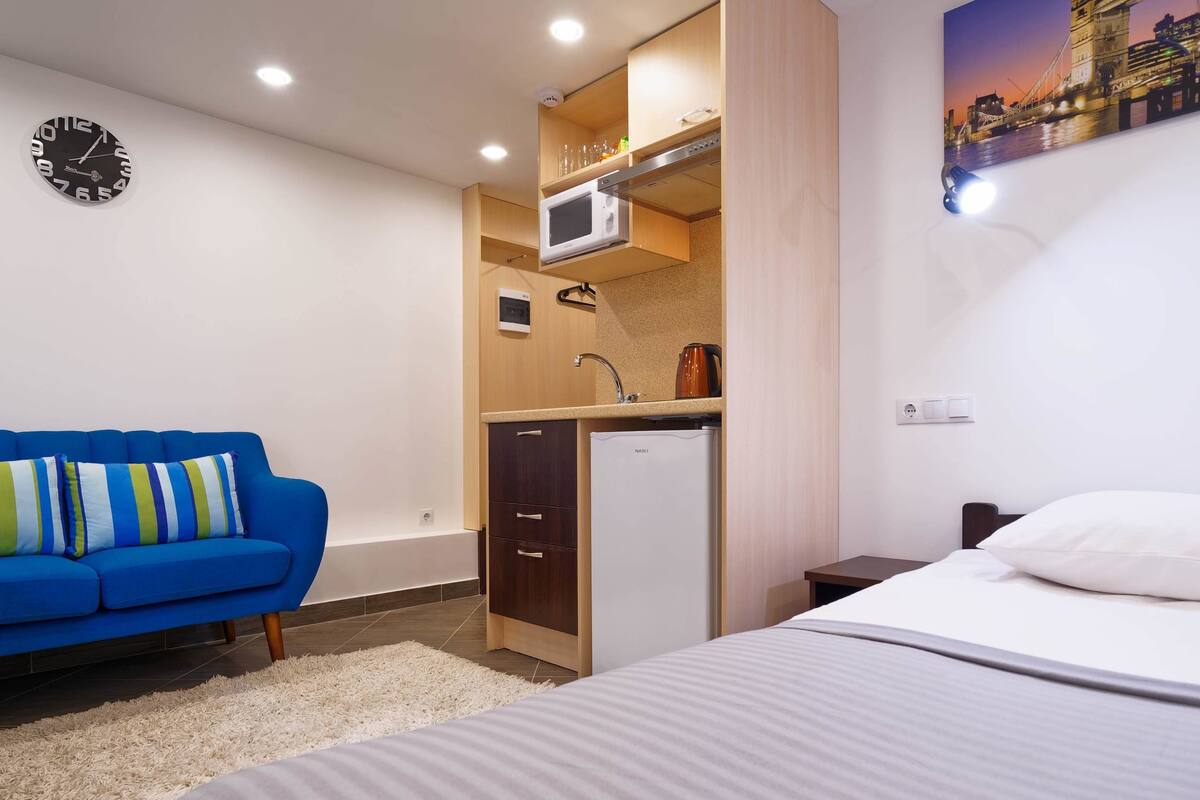 A compact hotel room with a bed, a blue sofa, a kitchenette with a microwave, and a clock on the wall.