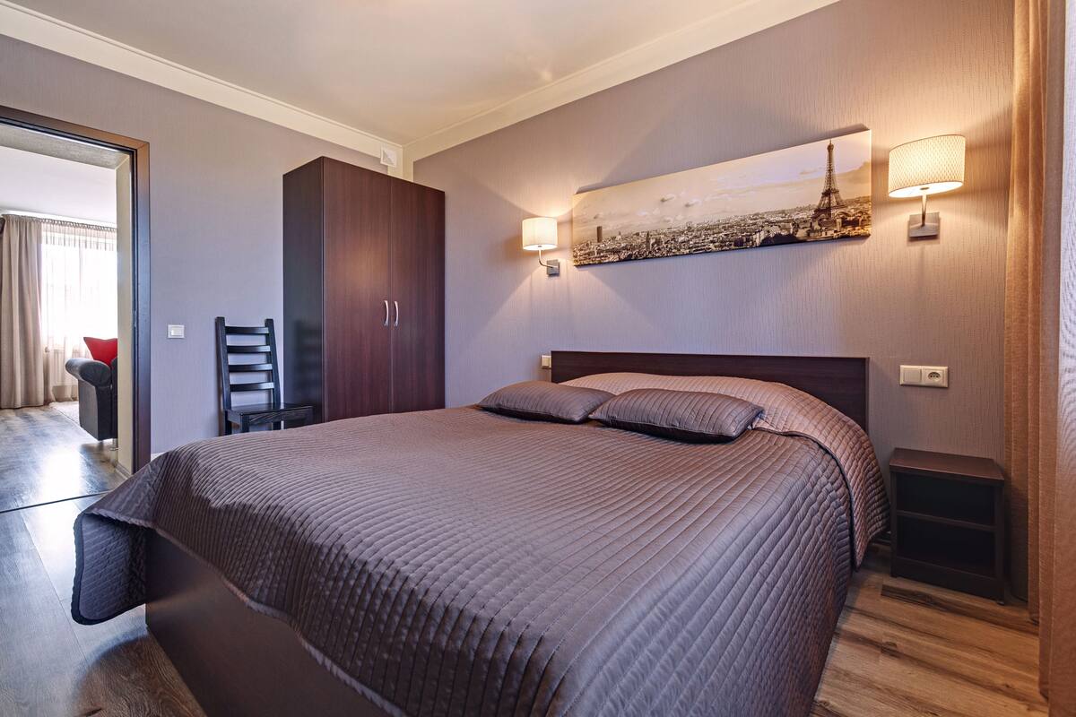 A neatly arranged bedroom with a large bed, a wardrobe, a chair, and a wall-mounted lamp.