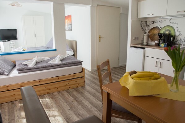A bedroom with a wooden bed, a bedside table, a chair, a small table with a banana bowl, and a kitchen area with a coffee maker and fruit. - Ferienwohnungen Meersburg (Meersburg)