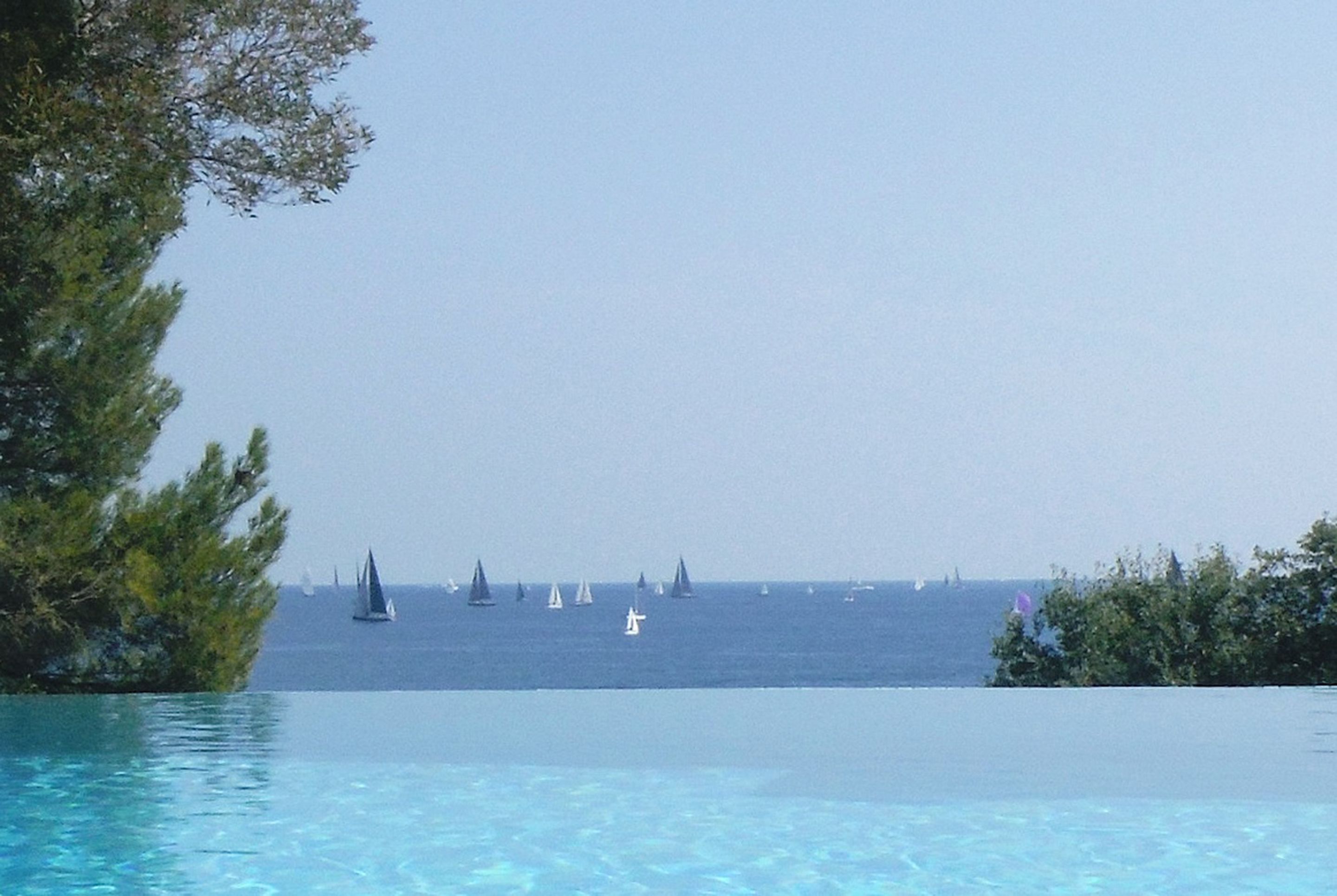 A pool with clear water, surrounded by greenery and a view of the sea with sailboats.
