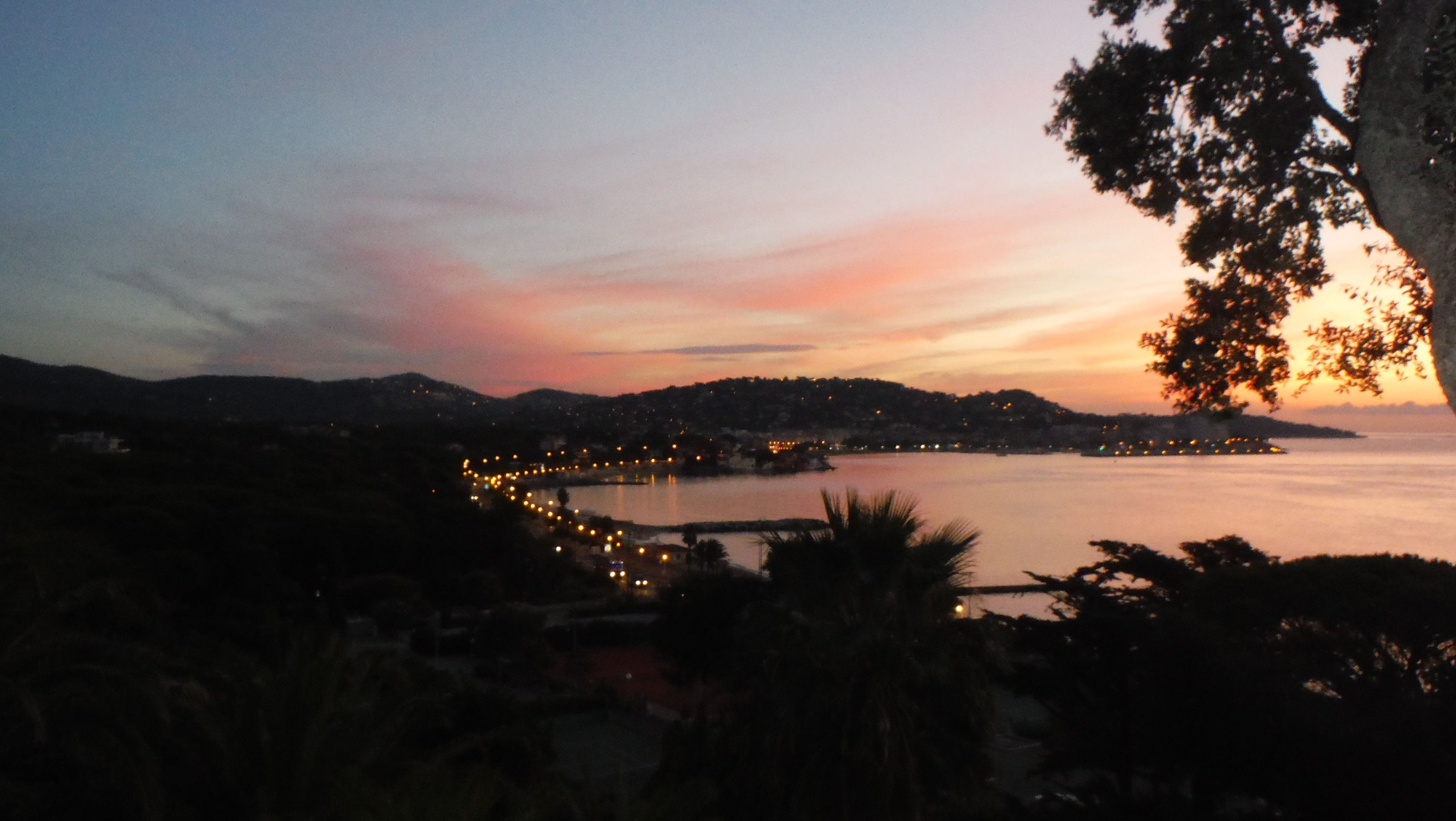 A coastal landscape at dusk with a view of a calm sea, a curved shoreline, and a distant cityscape with lights.