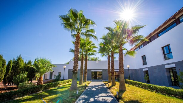 A modern building with a pathway lined by palm trees and a clear blue sky. - Hotel Las Monteras (Villanueva del Rey)