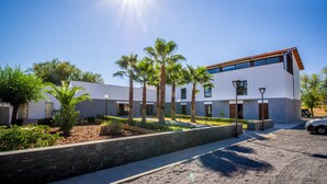 A modern building with a flat roof, large windows, and a paved courtyard with palm trees. - Hotel Las Monteras (Villanueva del Rey)