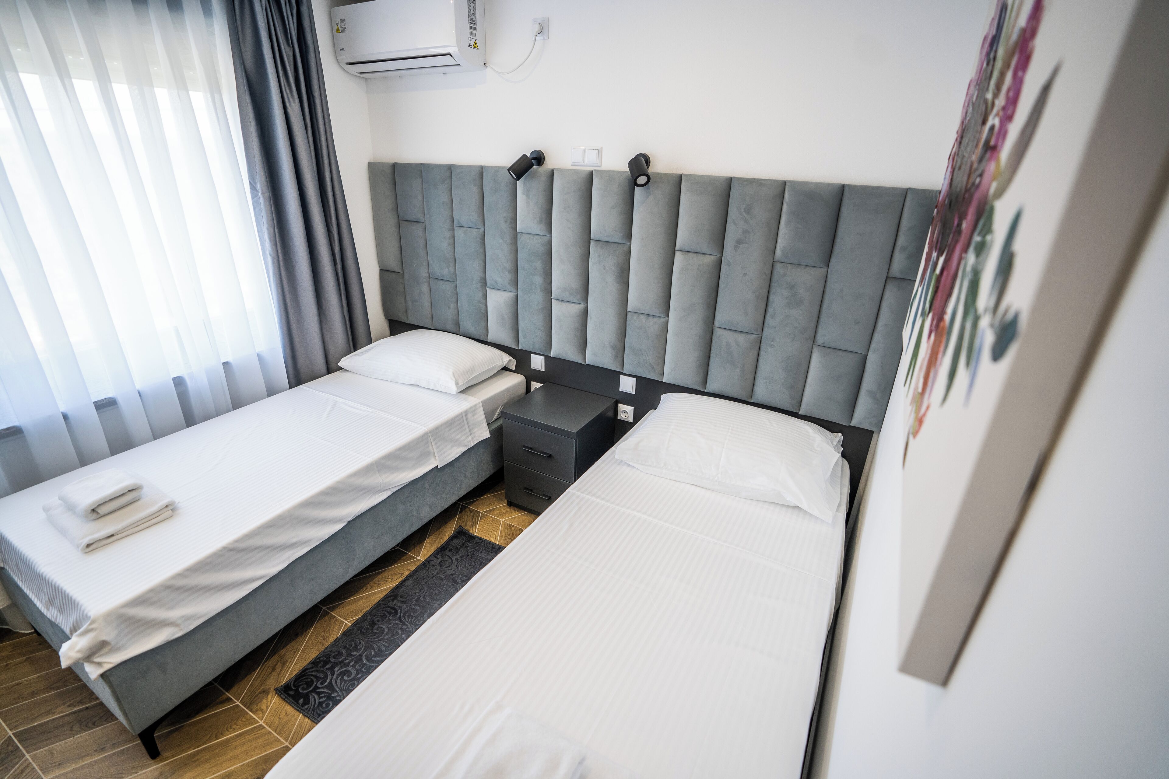 Habitación de hotel con dos camas individuales, cabecero gris, mesita de noche pequeña y aire acondicionado instalado en la pared.