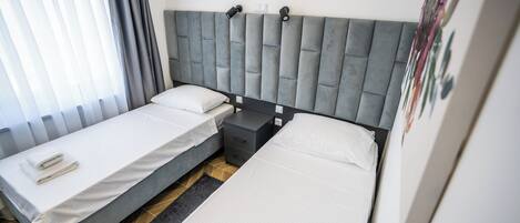 Habitación de hotel con dos camas individuales, cabecero gris, mesita de noche pequeña y aire acondicionado instalado en la pared.