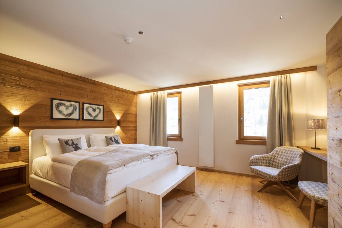 Un dormitorio moderno con techo de madera, una cama grande, un banco, una silla y dos ventanas con cortinas.