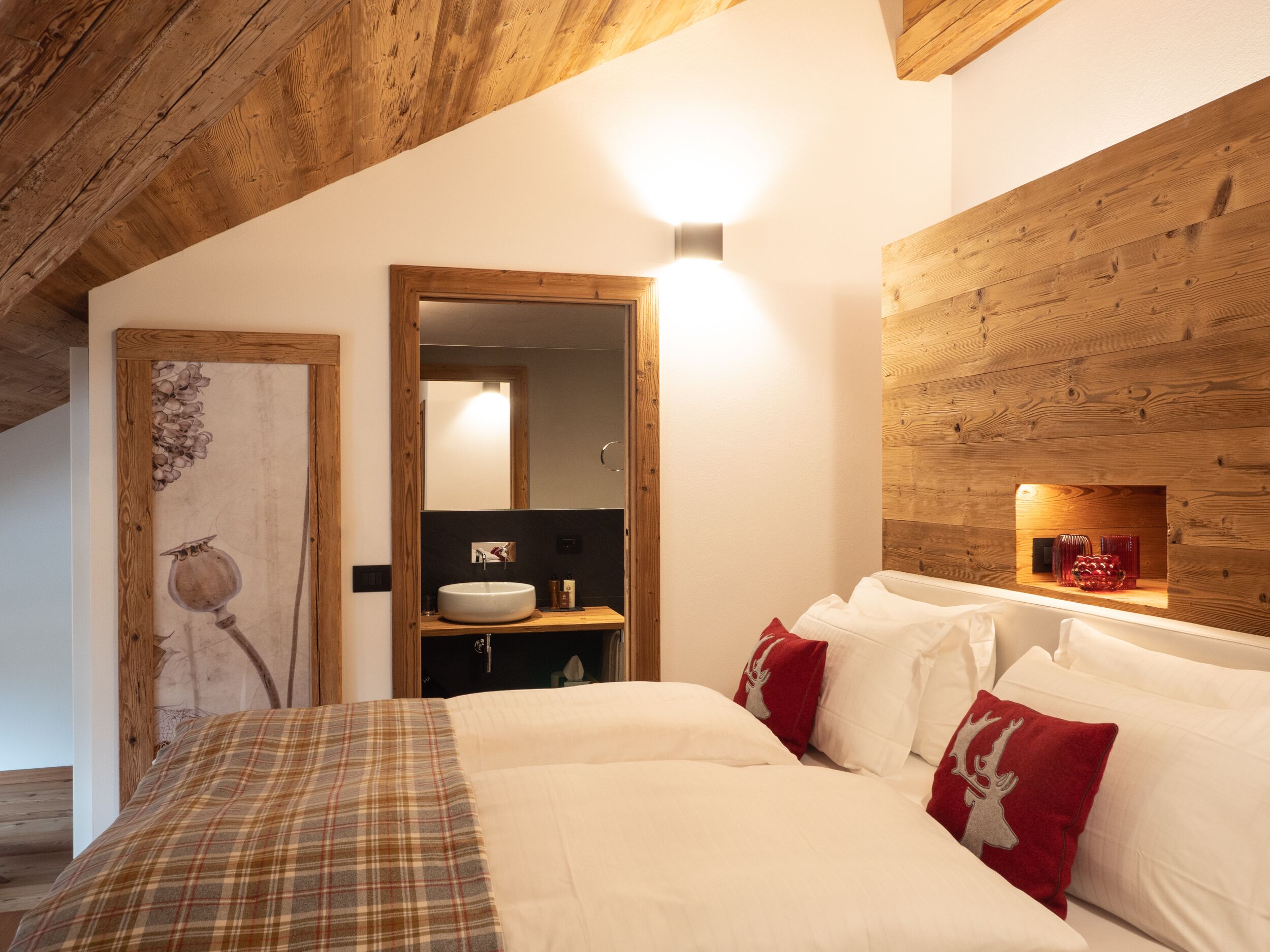 Una camera da letto con soffitto in legno, un letto grande con biancheria da letto bianca e cuscini rossi, uno specchio con cornice in legno e una parete in legno con uno scaffale.