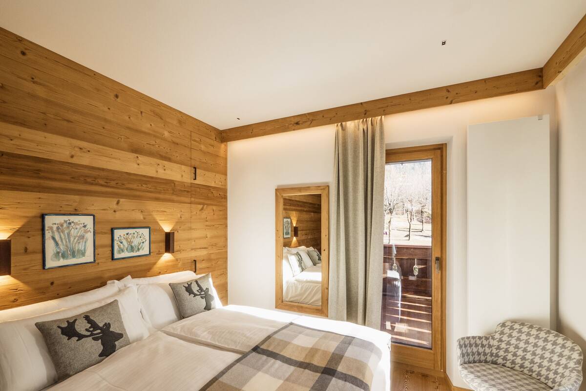 Un dormitorio con techo de madera, una cama grande con manta a cuadros, una silla y vistas a un balcón con barandilla.