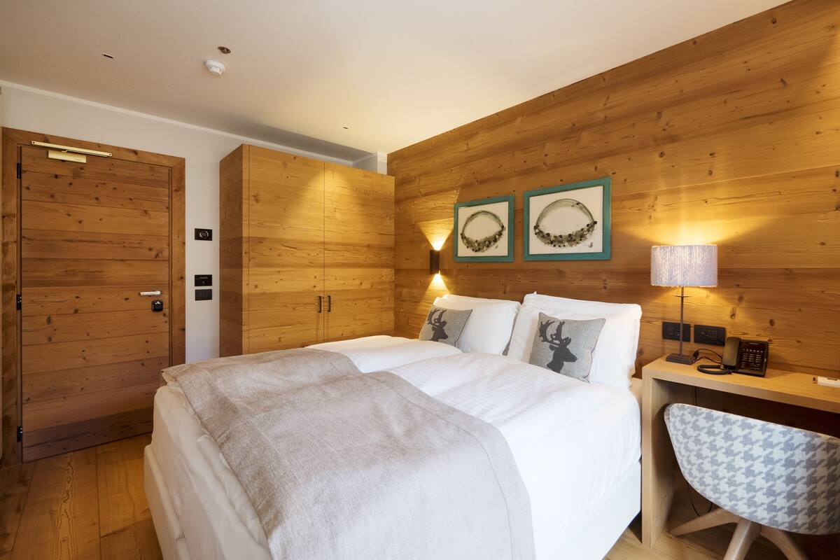 Un dormitorio con pared de madera, cama con ropa blanca, escritorio con lámpara y una silla.
