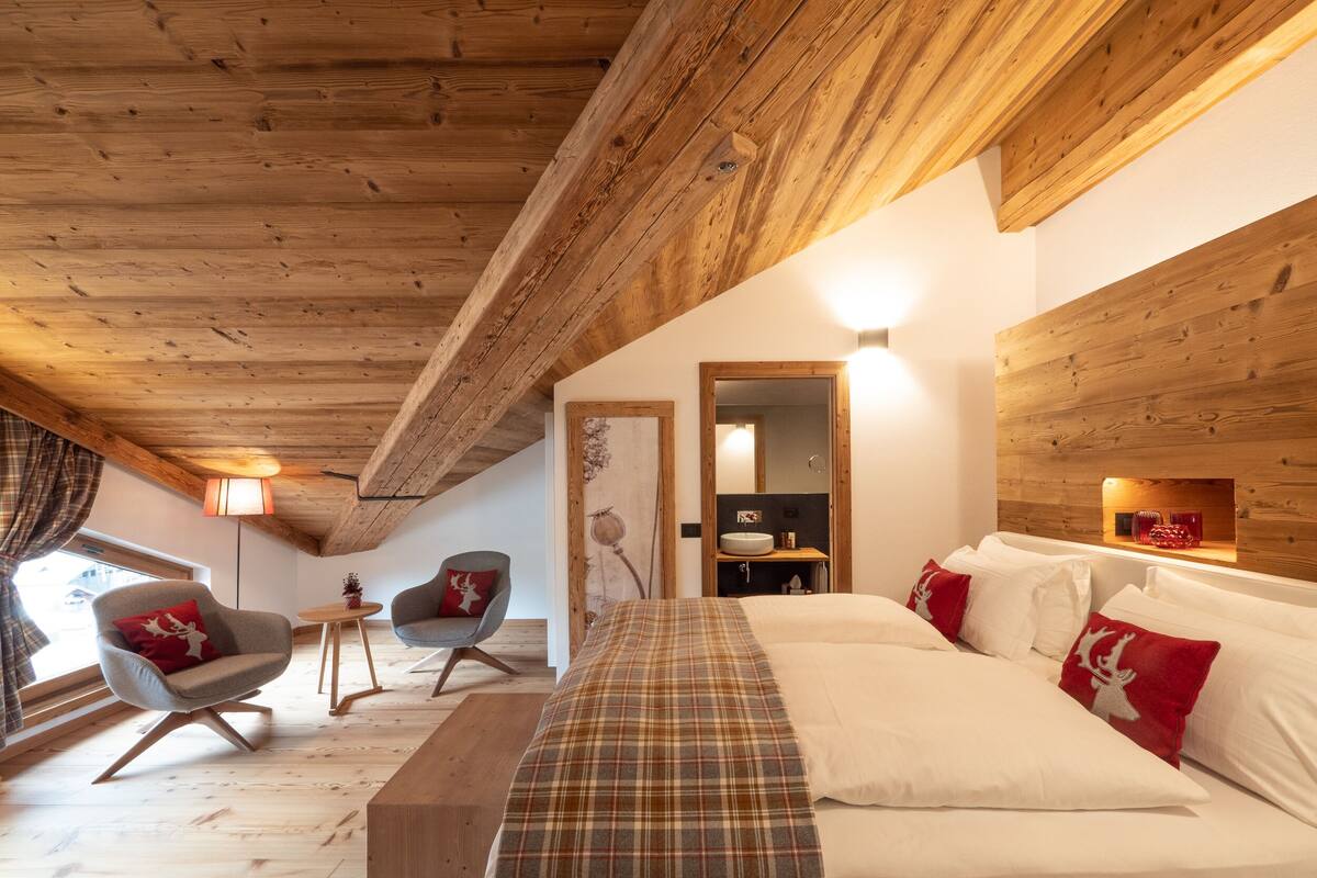 Un dormitorio acogedor con techo de madera, cama con ropa de cama escocesa, un banco y una silla con un cojín rojo.