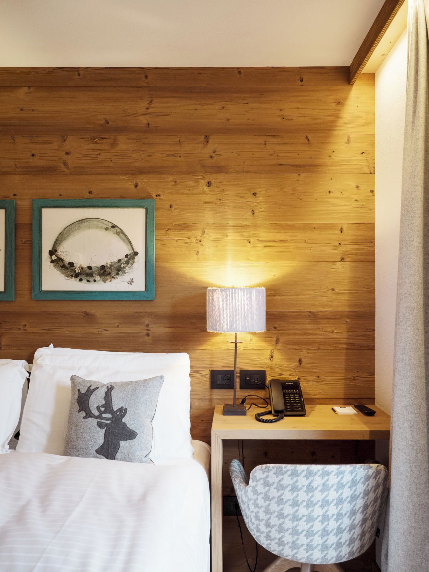 Habitación de hotel con pared de madera, cama con sábanas blancas, almohada gris con diseño de ciervo, escritorio pequeño con lámpara, teléfono y silla estampada.
