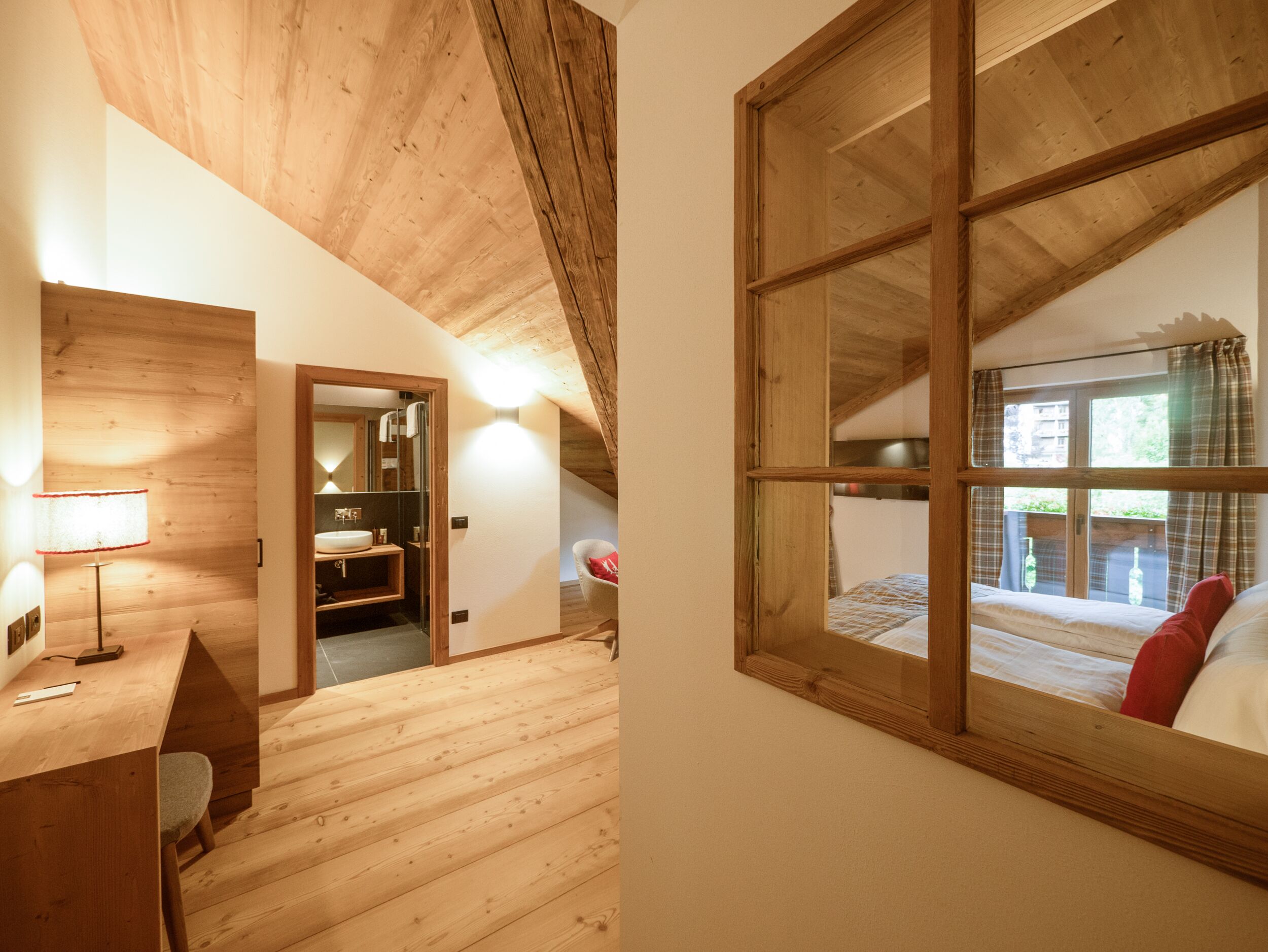 Una stanza con soffitto in legno, una scrivania con lampada, un letto con cuscino rosso e una finestra con tende.