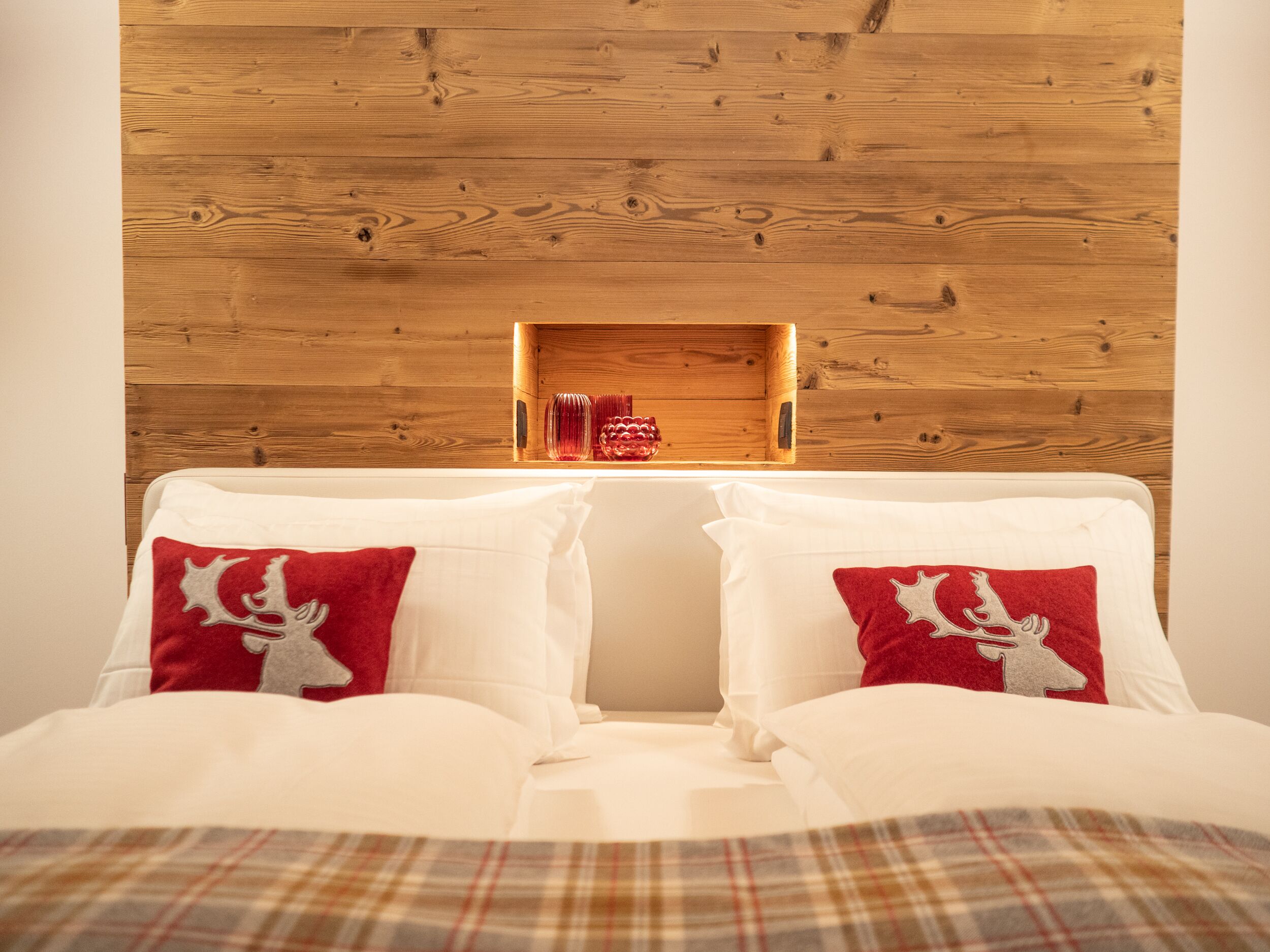 Uma cama com roupa de cama branca e almofadas vermelhas, cabeceira de madeira e uma pequena prateleira de madeira com objetos decorativos.