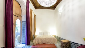 A narrow hallway leading to a bed with a patterned rug, a wooden door with intricate carvings, and a ceiling with a decorative light fixture. - Riad La maison d'à côté (Meknes)