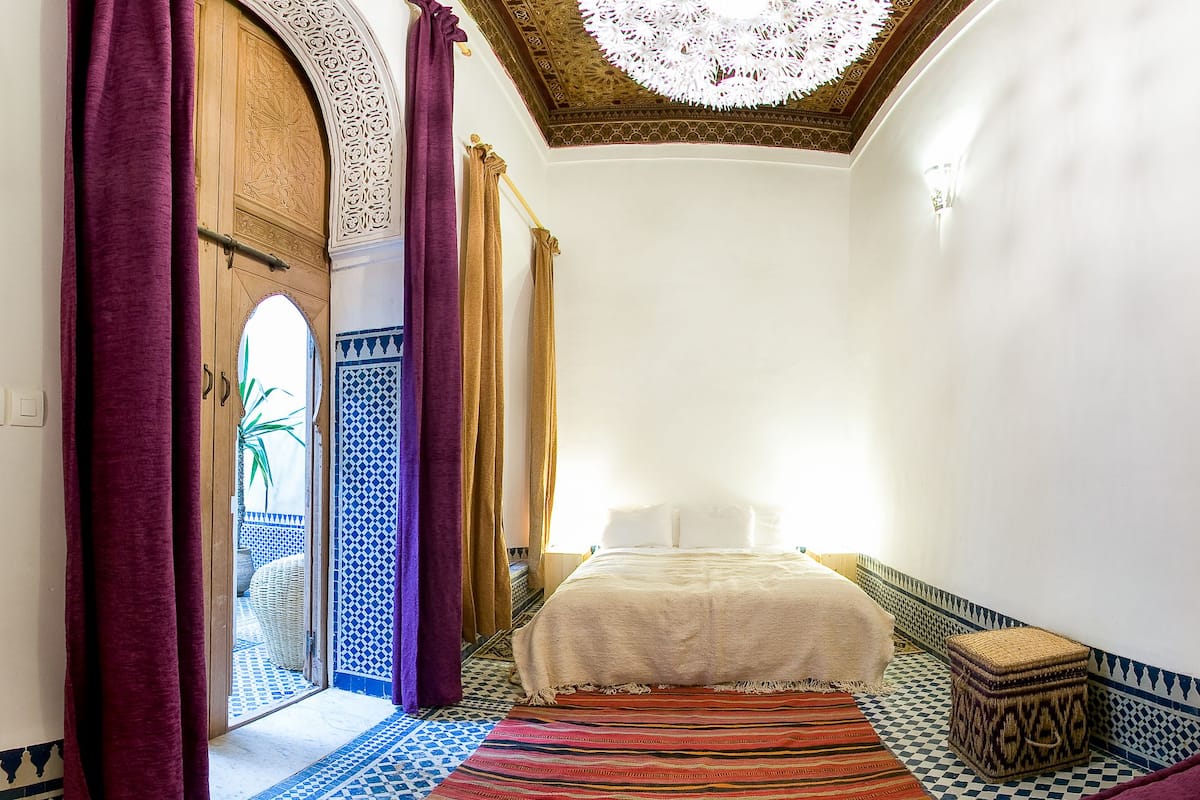 Un pasillo estrecho que conduce a una cama con un tapete estampado, una puerta de madera con intrincados relieves y un techo con una lámpara decorativa.