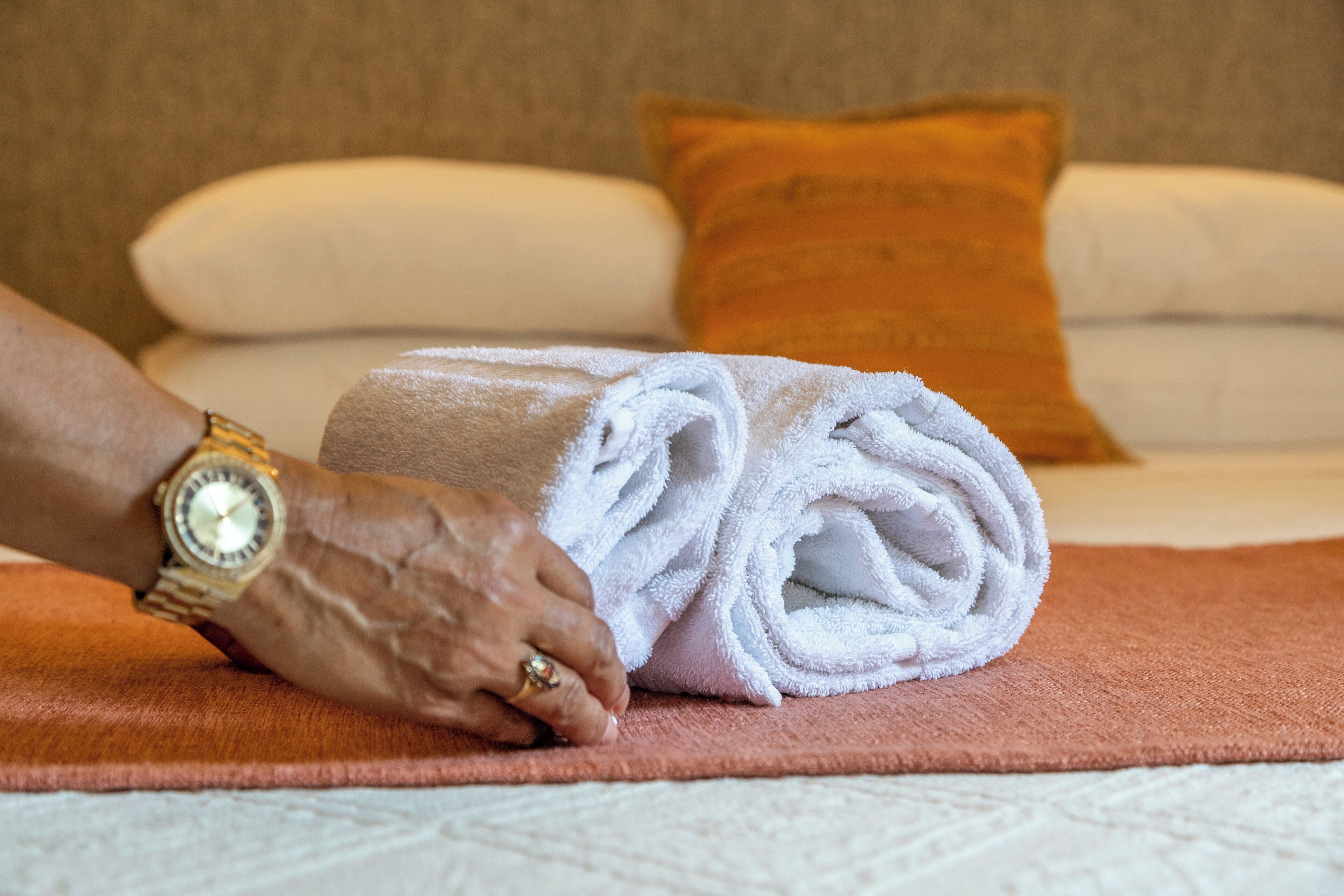 Una toalla doblada cuidadosamente sobre un tapete marrón, junto a una cama con almohadas blancas y anaranjadas.