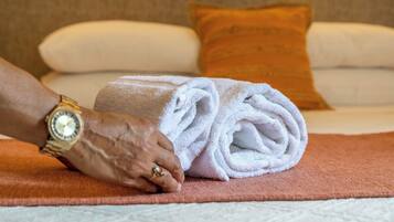 Una toalla doblada cuidadosamente sobre un tapete marrón, junto a una cama con almohadas blancas y anaranjadas.