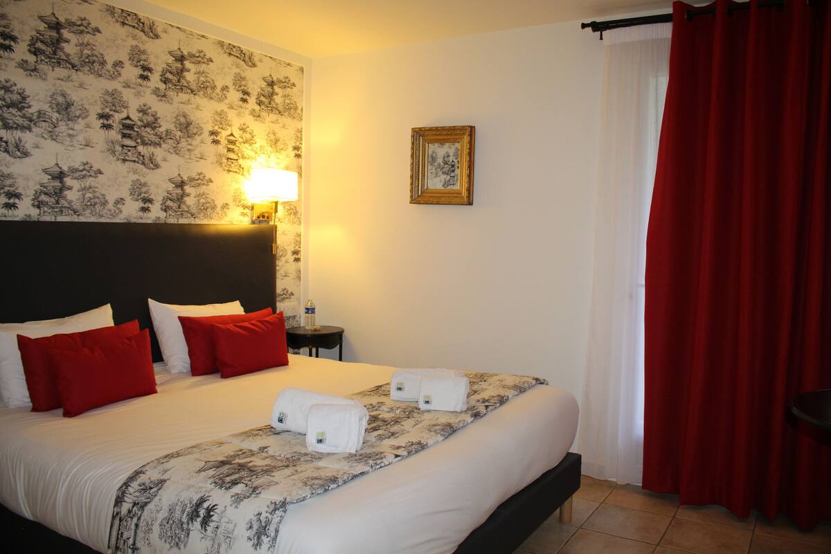 A hotel room with a large bed, red and white pillows, a headboard, a wall-mounted lamp, a framed picture, and a red curtain.