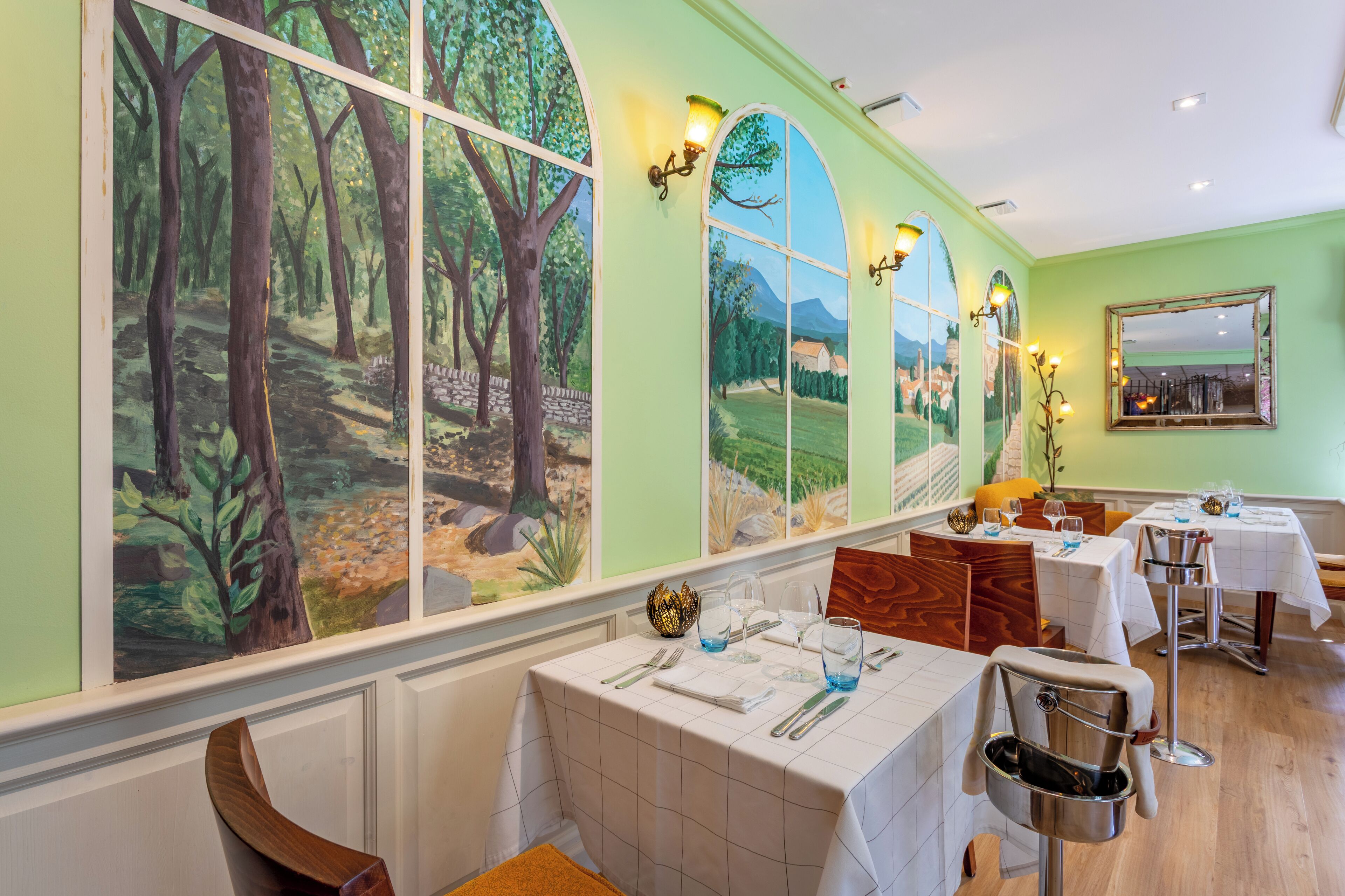 A restaurant with a forest mural, white tablecloths, and wooden chairs.