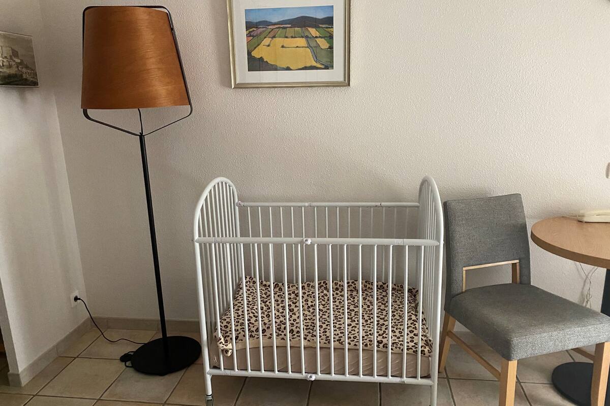 A white crib with a leopard print mattress, a grey chair, a floor lamp, and a framed picture on the wall.
