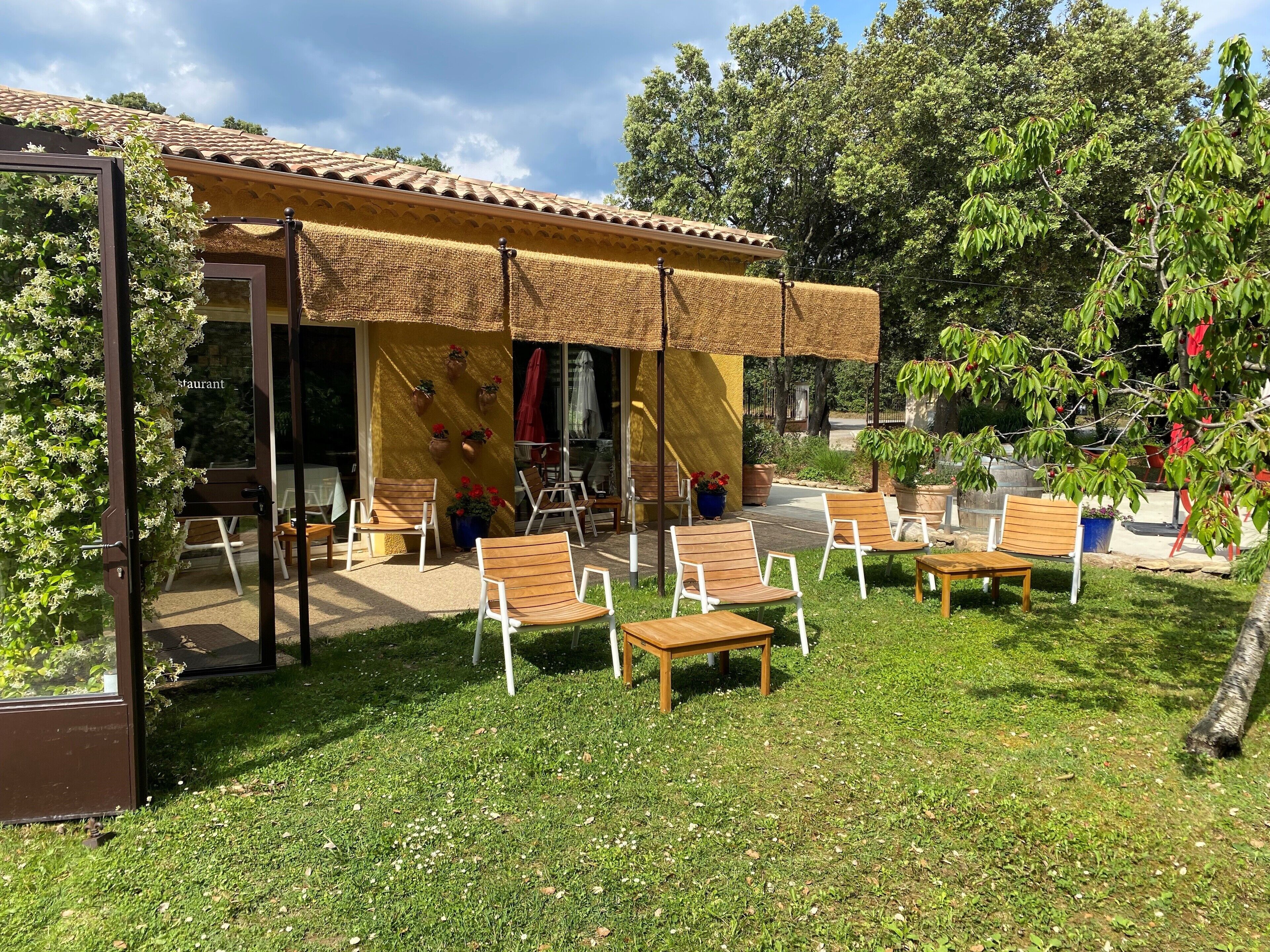 An outdoor seating area with wooden chairs and tables, a yellow building with a tiled roof, and potted plants.