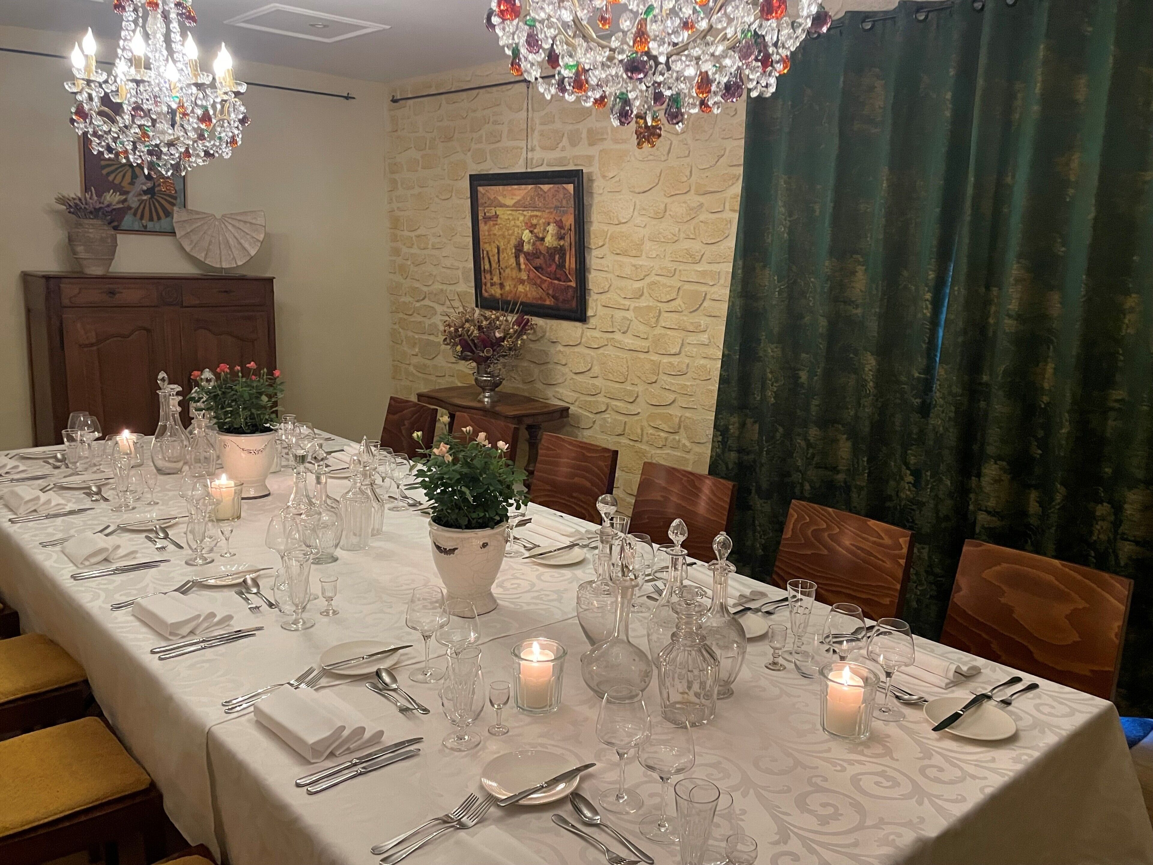 A dining table set with white tablecloth, candles, and glassware, with a chandelier and a painting on the wall.