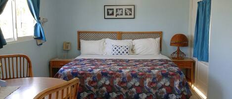 Un dormitorio con cama, cabecera de madera, mesita de noche con lámpara, una silla y ventana con cortinas.