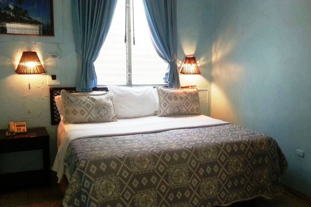 A bedroom with a bed, two bedside lamps, a nightstand, and a window with curtains.