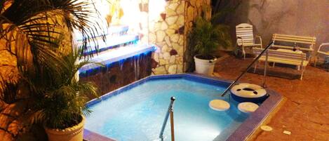 A small outdoor swimming pool with a stone wall, a hot tub, and a few lounge chairs.
