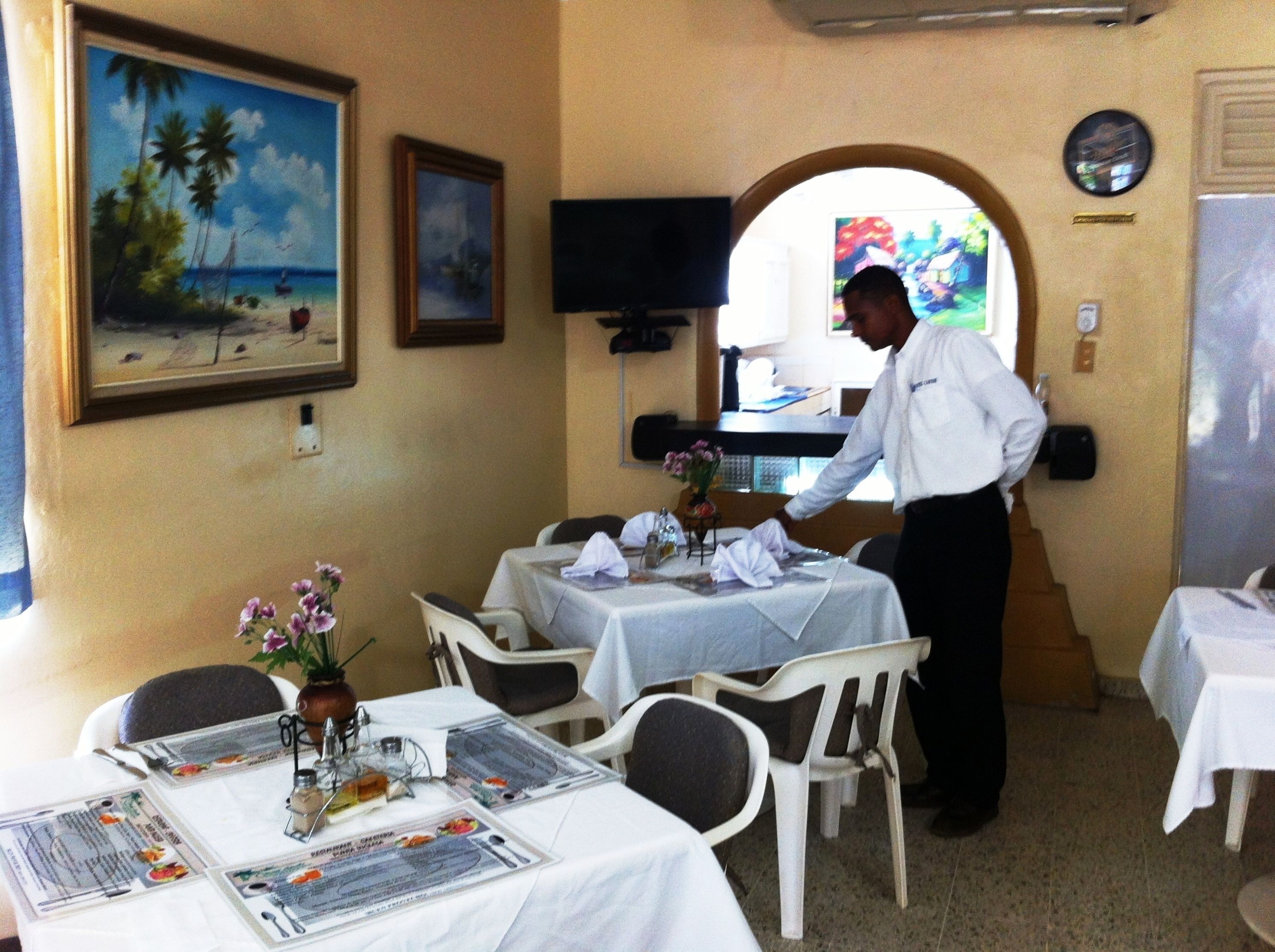 A restaurant with tables set for dining, a waiter attending to guests, and a painting of a beach scene on the wall.