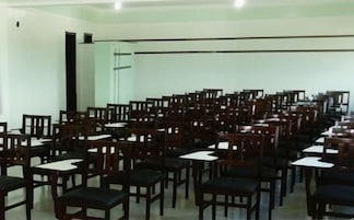 Uma sala de conferências com fileiras de cadeiras e mesas de madeira, paredes brancas e uma janela grande.