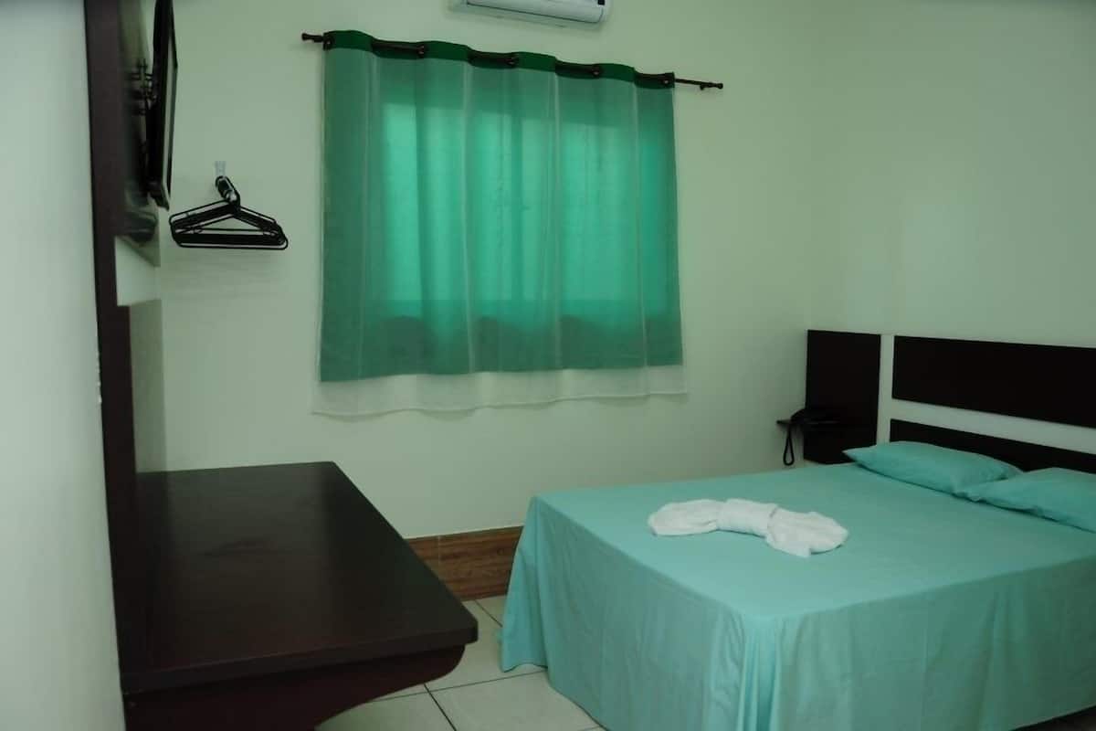A single bed with a teal bedspread and a white towel, a bedside table, a wall-mounted telephone, and a green curtain.