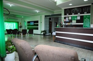 Zona de recepción de hotel con mostrador, varias sillas, un televisor y una pared verde con relojes.