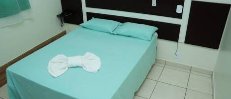 Una cama individual con una colcha verde azulada y almohadas blancas, un cabecero negro, un teléfono en la mesita de noche y una toalla blanca doblada sobre la cama.