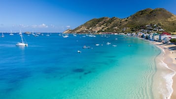 A coastal area with clear turquoise waters, boats, and a hilly backdrop with buildings.