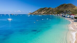 A coastal area with clear turquoise waters, boats, and a hilly backdrop with buildings. - Le Temps des Cerises Hotel Beach (Grand Case)