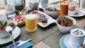 Une table de petit-déjeuner garnie de divers aliments et boissons, donnant sur une plage où l’on peut voir des voiliers.