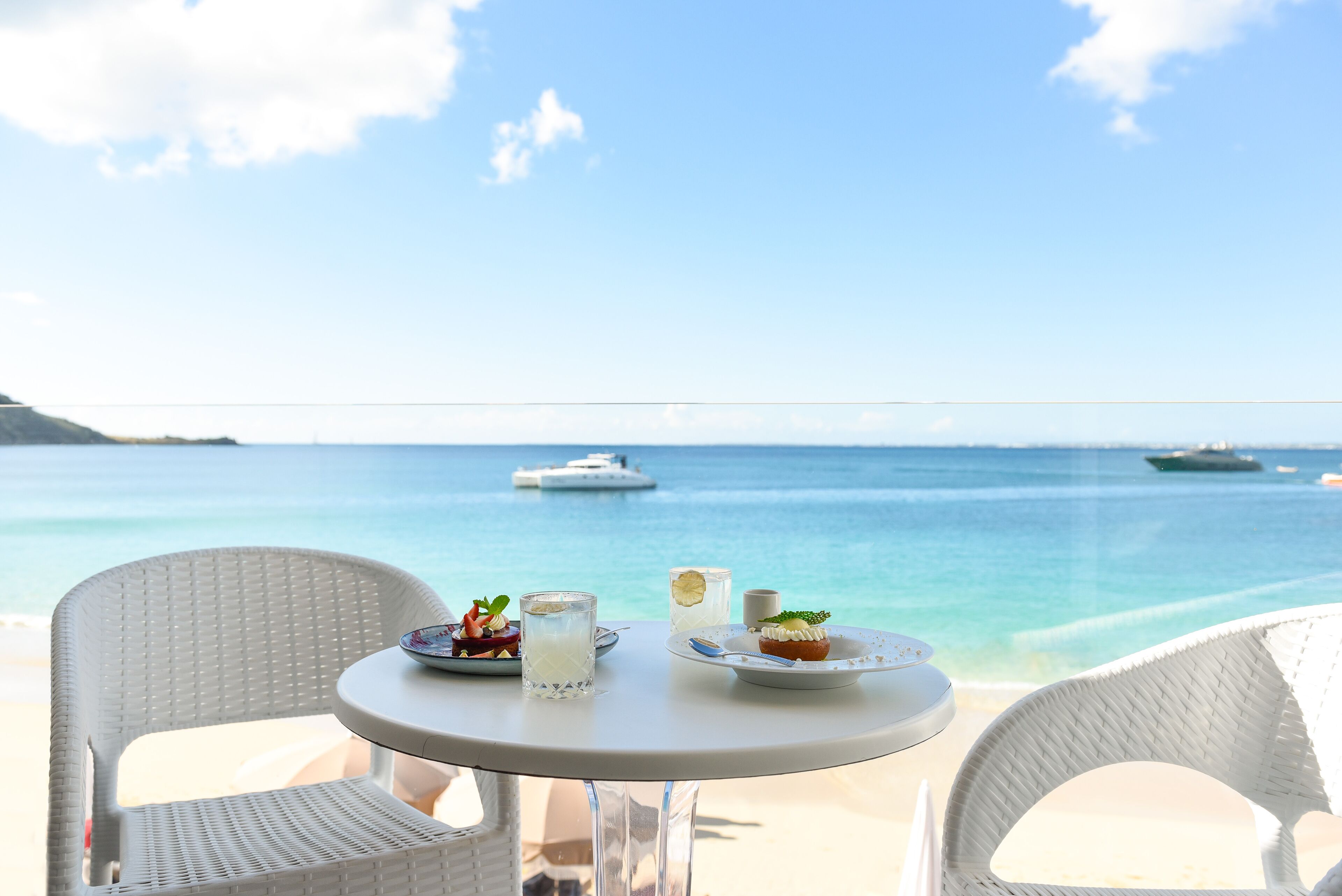 Una mesa con platos de comida y bebidas, con vista a una playa donde se ven barcos a lo lejos.