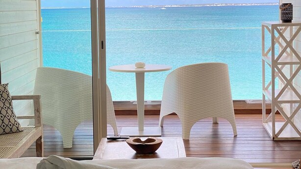 Un balcon avec vue sur la mer, un lit avec des serviettes et deux chaises en osier blanches.