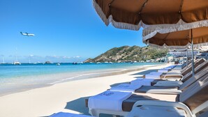 Une plage bordée de rangées de transats sous des parasols, un ciel bleu azur et un avion qui vole au loin.