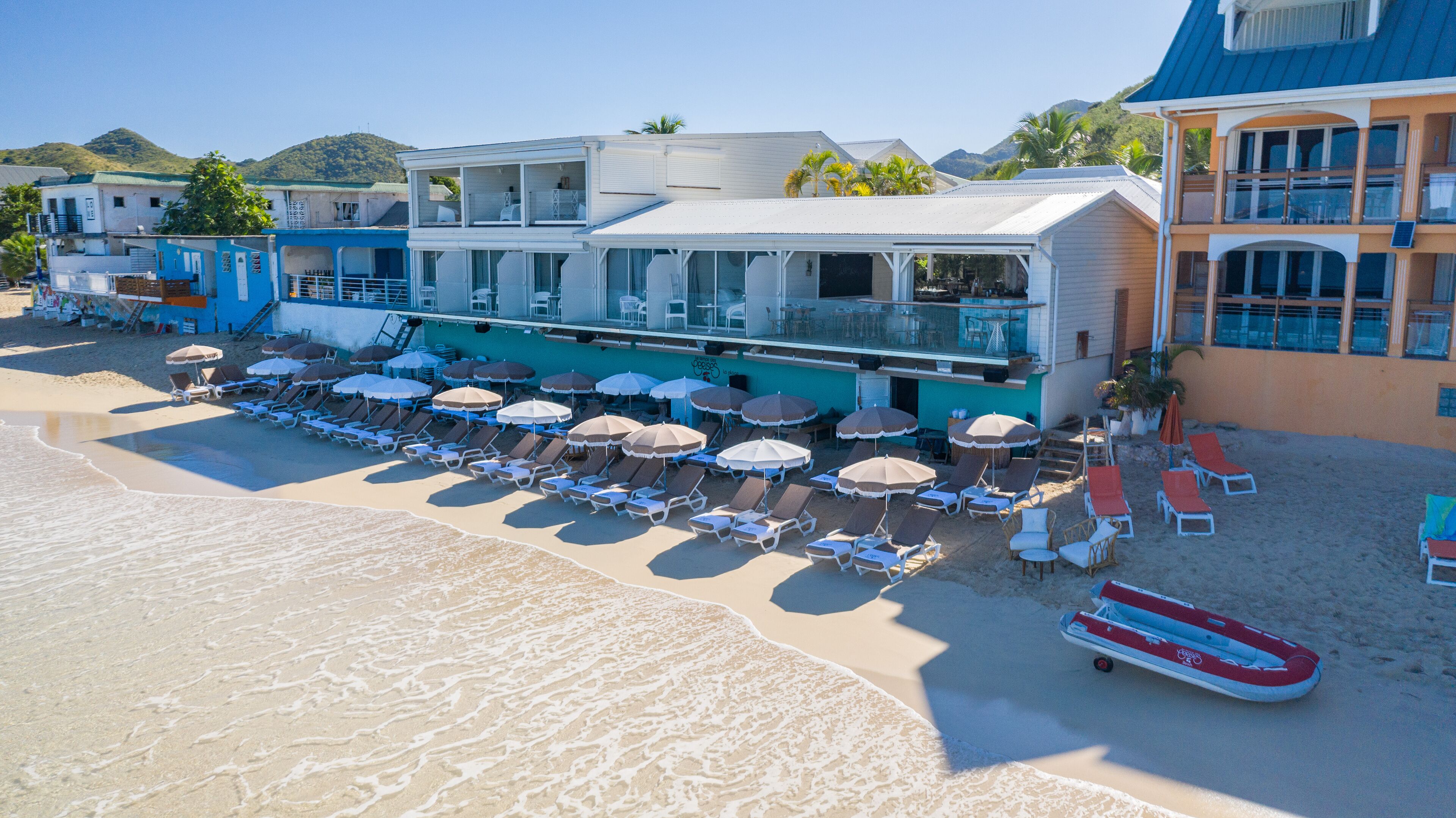 Un hôtel situé en bord de mer, doté de plusieurs espaces de détente et de parasols, d’un bateau de sauveteurs, et offrant une vue sur l’océan et les collines en arrière-plan.