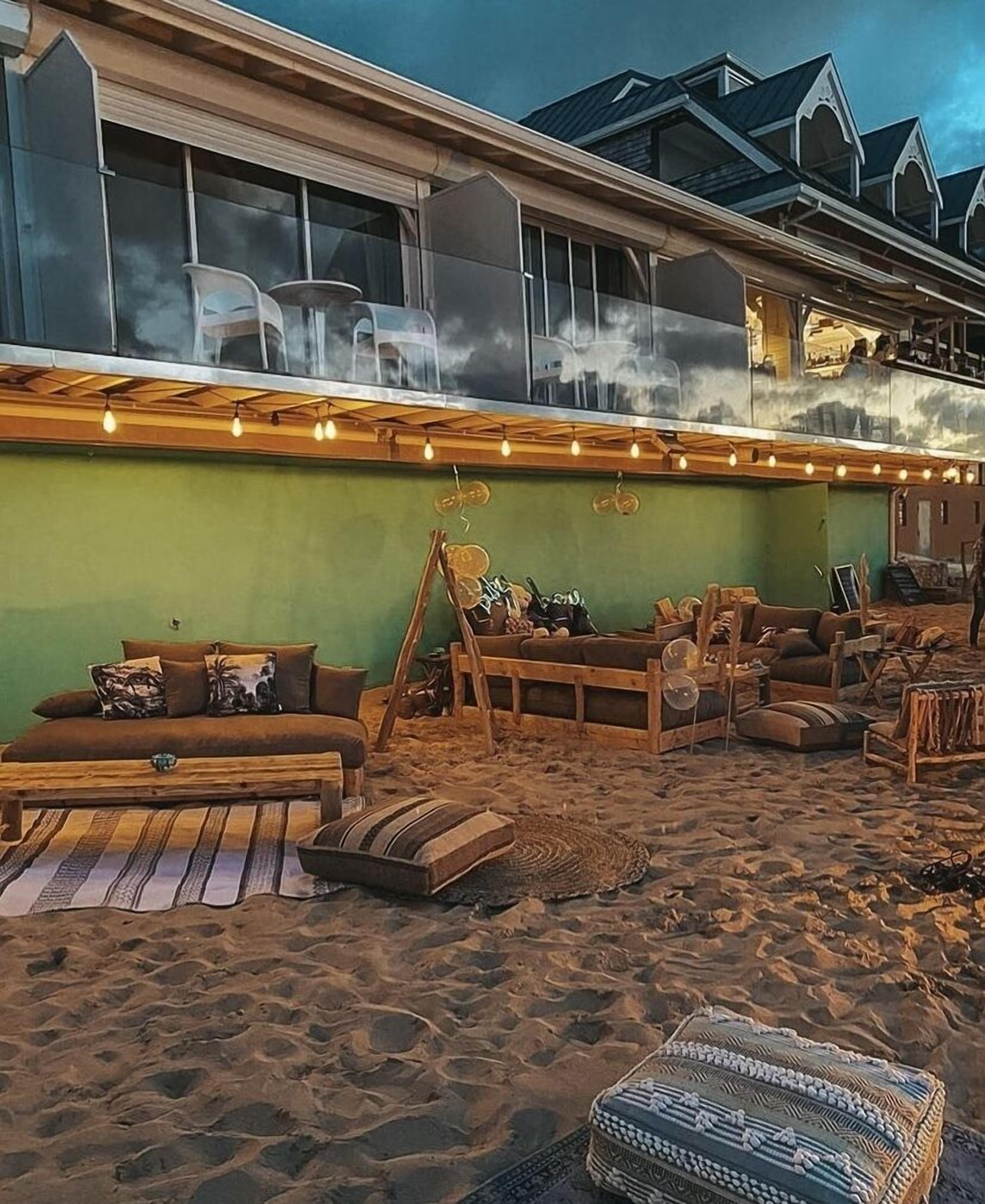 A beachside lounge with wooden furniture, a green wall, and string lights.